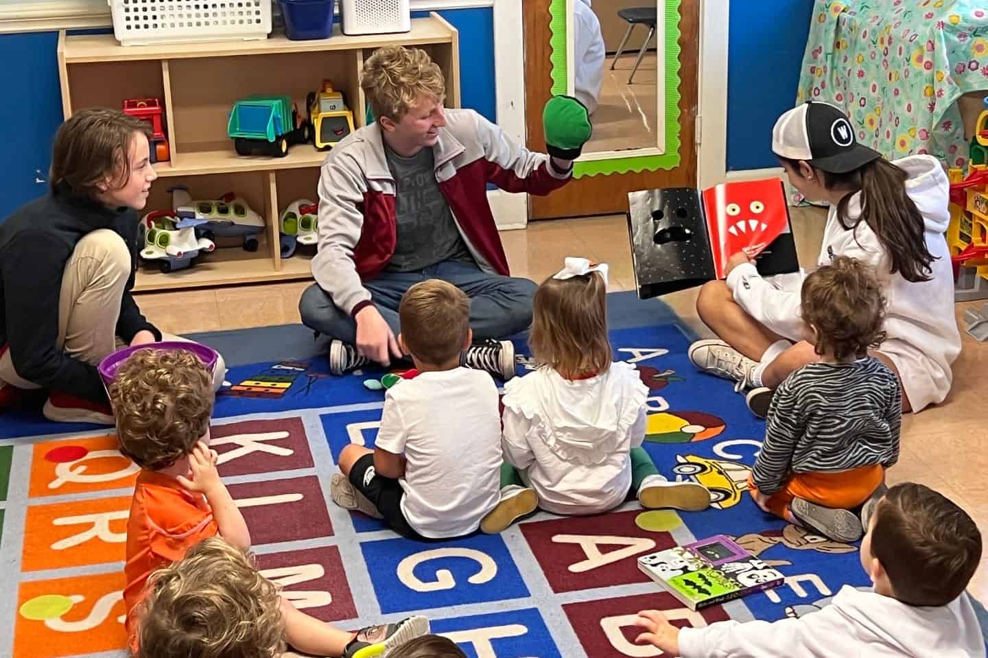 An older student holds a puppet while another older student readers a book to a group of young children