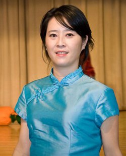 A smiling woman, Wei Ma, pictured indoors in front of a stage curtain, looking at the camera