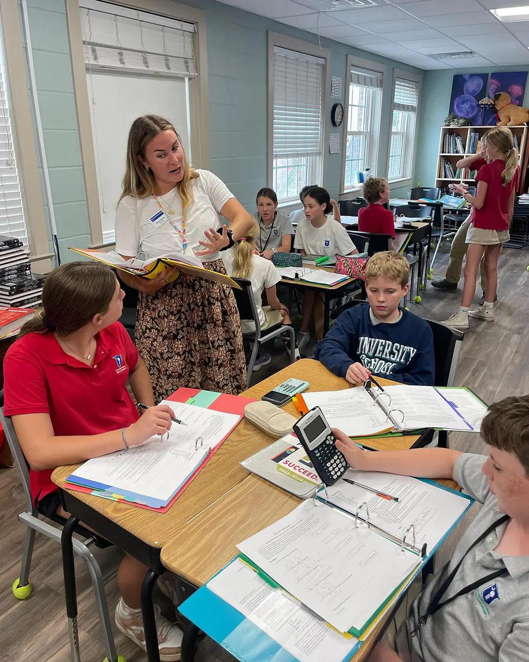 In a classroom setting, a teacher stands over a student, explaining a concept, while other students surround them. Students work in small groups in the background.