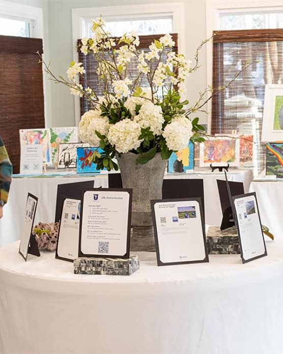 A round table covered with a white tablecloth displays framed auction item descriptions on black stands, with QR codes visible on the printed sheets. A tall centerpiece of white hydrangeas and flowering branches in a rustic metal vase decorates the center of the table. In the background, colorful artwork and additional auction items are visible, set up on tables near large windows with wooden blinds. The setting appears to be a bright, well-lit indoor event space.